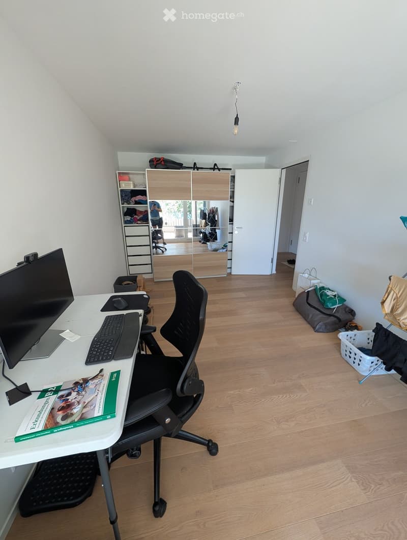 A well-lit room with wooden flooring, a desk with a computer setup, a chair, a wardrobe with a mirror, and various storage items.