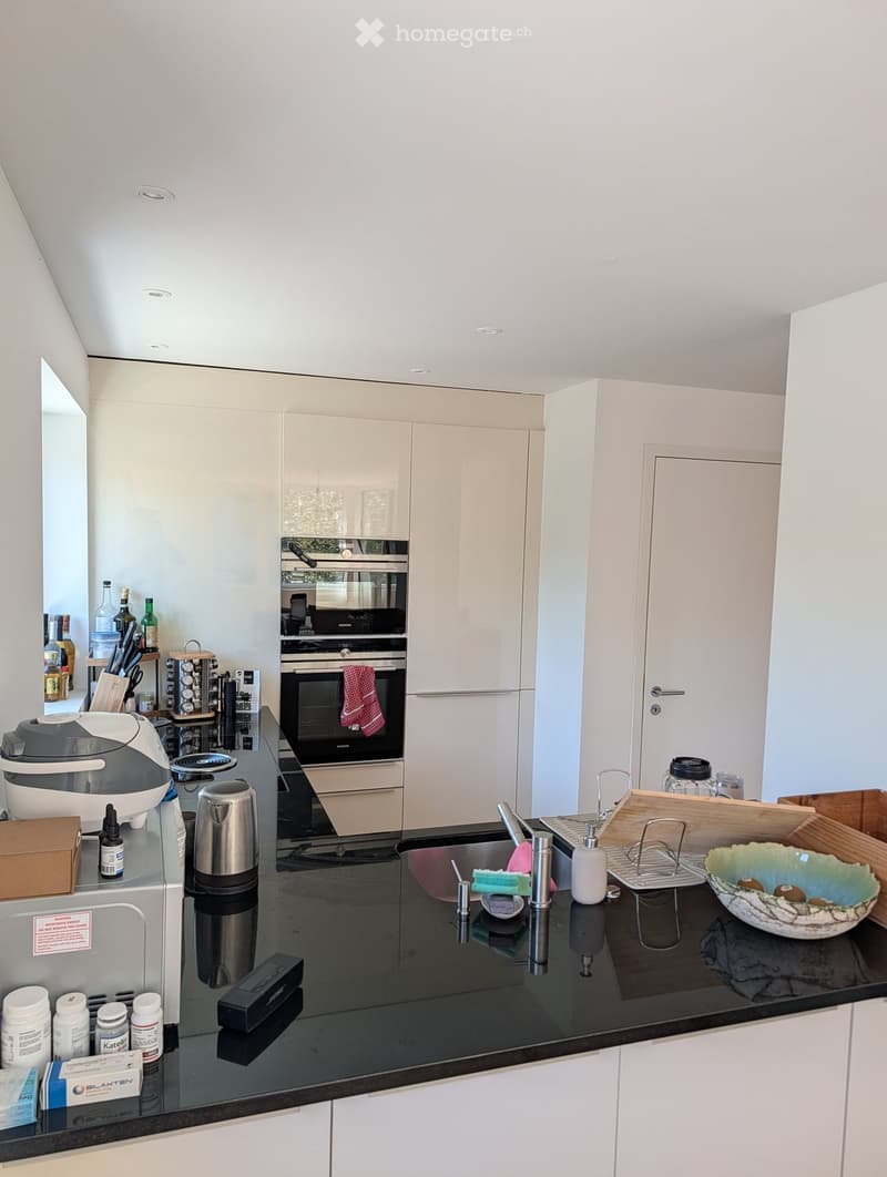 Modern kitchen with white cabinets, black countertop, stainless steel appliances, and various kitchen items including a coffee maker, bottles, and a bowl on the counter.