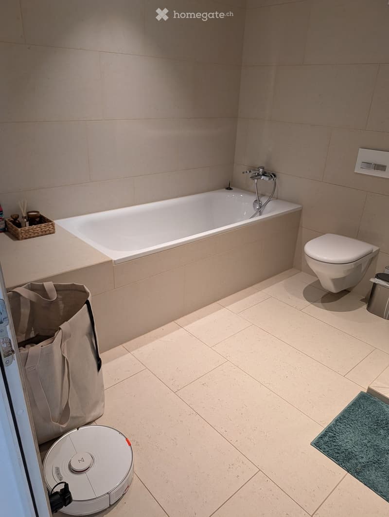 Modern bathroom with a bathtub, toilet, and a vacuum cleaner on the floor. Beige tiles and a small green rug are visible.