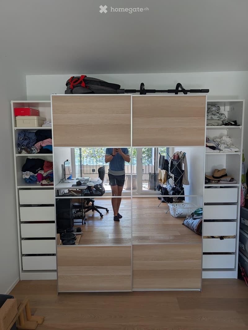 A modern, well-organized room with wooden flooring, large sliding doors leading to a balcony, and multiple storage units including shelves, drawers, and a clothing rack. A desk with a chair and laptop is visible, along with various personal items such as