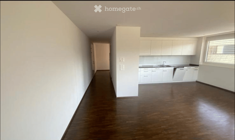 Empty hallway leading to a kitchen area with white cabinets and stainless steel appliances, featuring dark hardwood flooring and white walls.