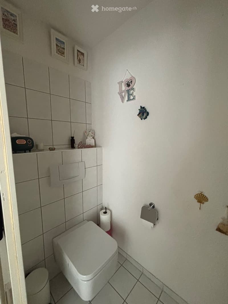 A small bathroom with a toilet, toilet paper holder, and decorative wall art including a 'LOVE' sign and framed pictures. The walls are tiled, and there is a radiator on the left side.