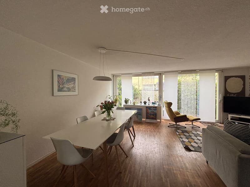 Modern living and dining area with wooden flooring, large windows, a sofa, a dining table with chairs, a television, and decorative items.
