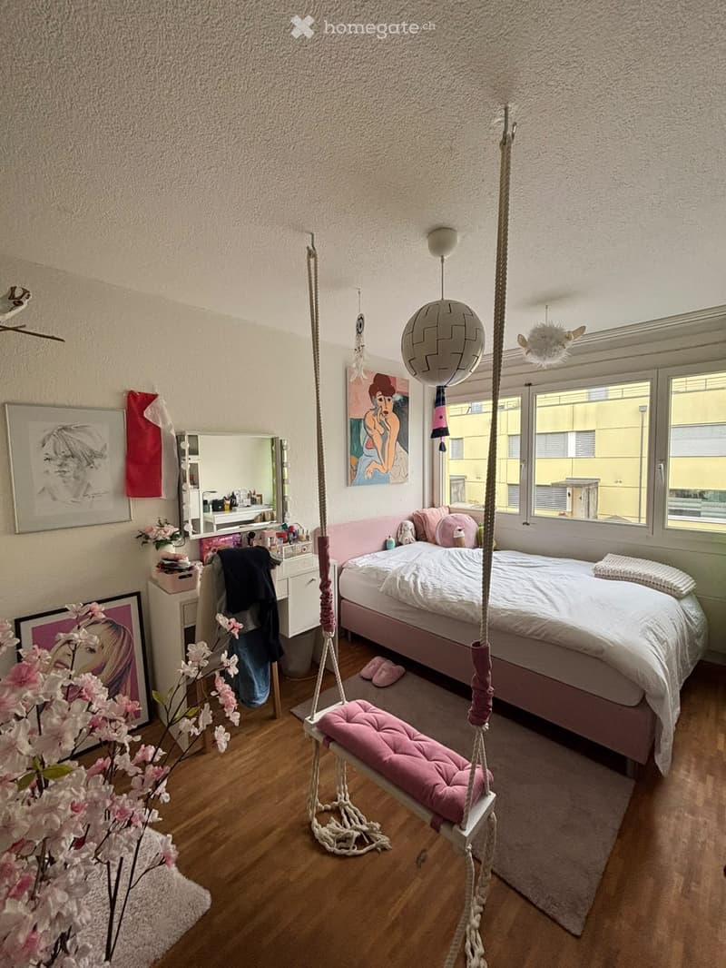 Cozy bedroom with a pink bed, white bedding, a wicker hanging chair, framed artwork, a mirror, and a flower arrangement on the floor.