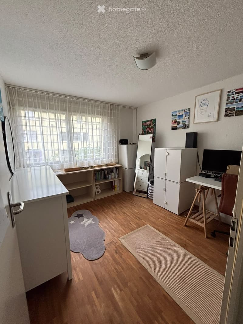 A well-lit room with wooden flooring, a desk with a chair, a cabinet, a mirror, a refrigerator, a shelf with books, a speaker, a computer monitor, a keyboard, picture frames, a curtain, and rugs.
