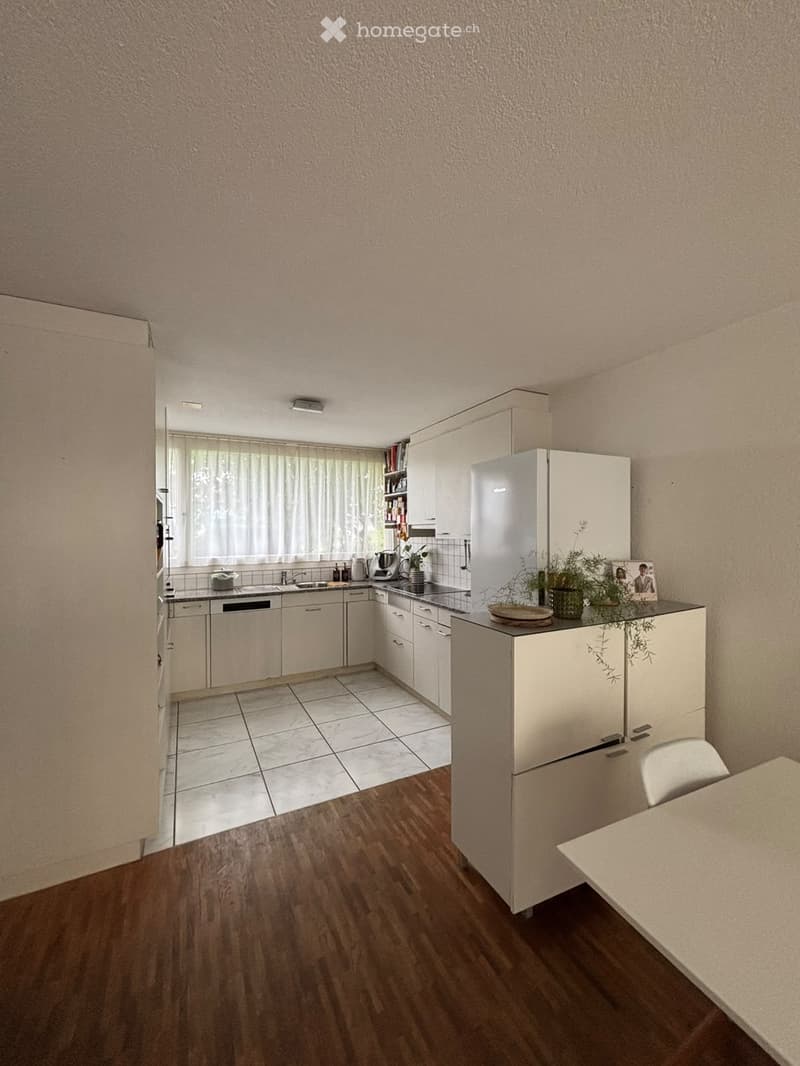 Modern kitchen with white cabinets, a refrigerator, a sink, a dishwasher, and a small dining area with a table and chair. The floor is a combination of tiled and wooden surfaces, and there is a window with a curtain.