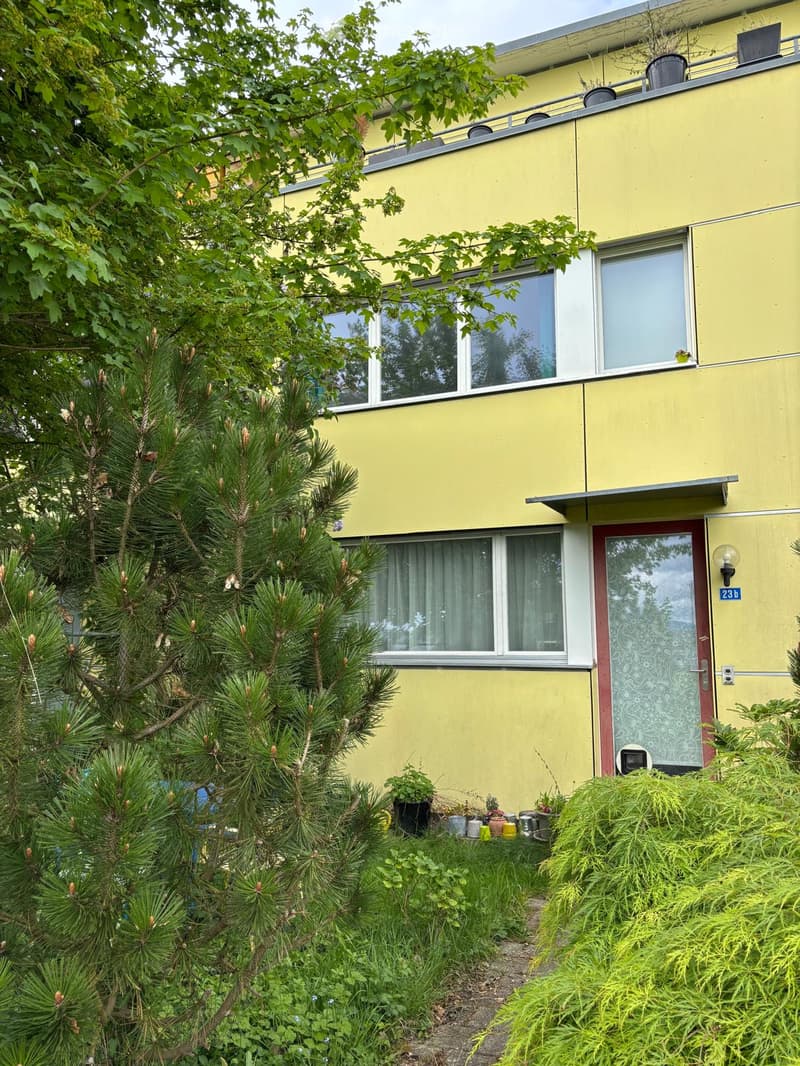 Two-story yellow building with white trim, multiple windows, and a small balcony on the upper floor. The entrance has a red door with a decorative glass panel. The building is surrounded by greenery, including a pine tree and various plants.