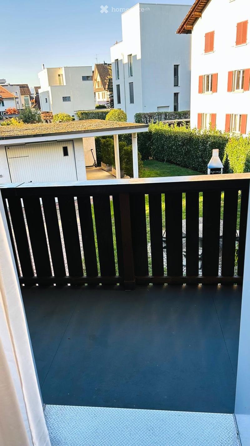 Balcony with dark wooden railing, grey tiled floor, and a view of a residential area with houses, greenery, and a garage.