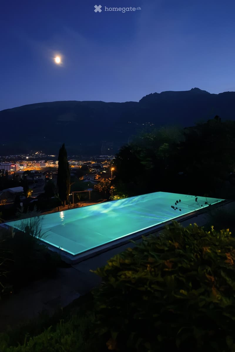 A modern swimming pool with a sleek, illuminated design, surrounded by lush greenery and overlooking a cityscape and mountains at night.