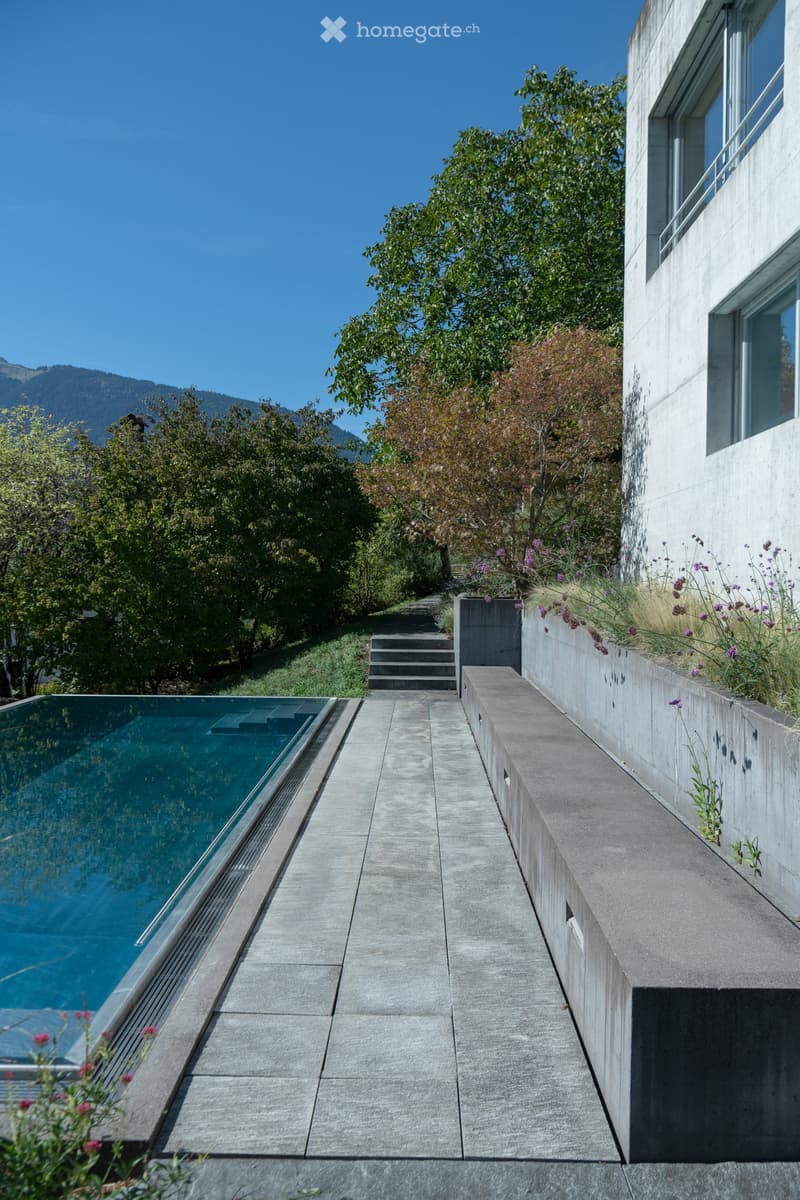 Modern outdoor area with a sleek swimming pool, concrete pathways, steps, and surrounding greenery, featuring a raised ground floor and scenic mountain view.