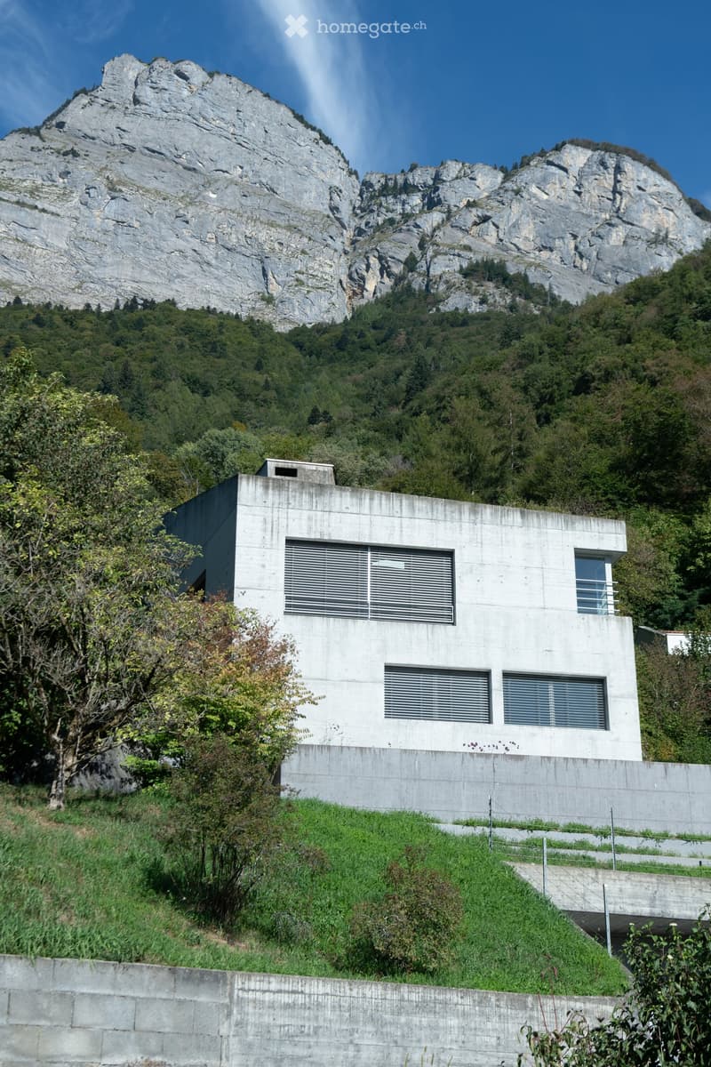 Modern concrete building with large windows and blinds, situated in a lush, green landscape with a prominent rocky mountain in the background.