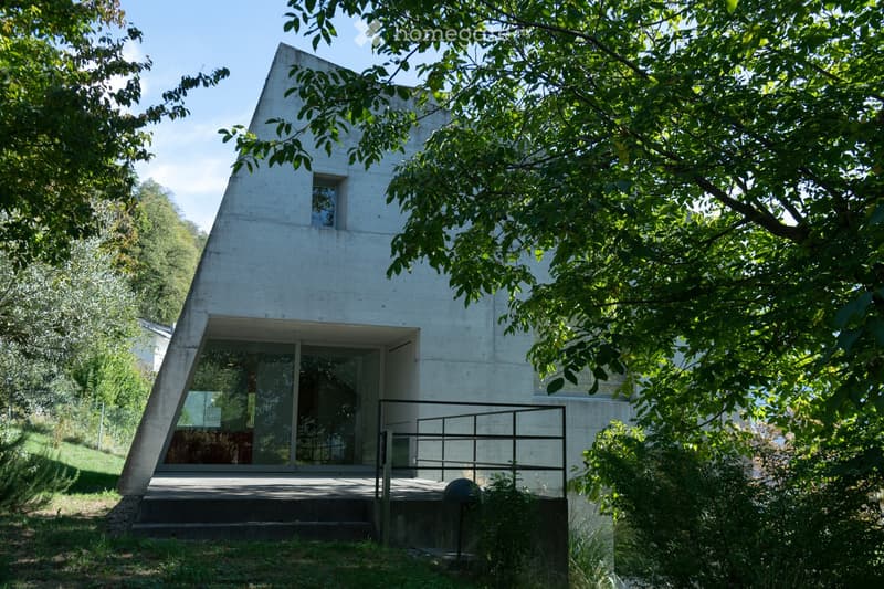 Modern, minimalist house with a concrete facade, large glass windows, and a raised ground floor surrounded by lush greenery.