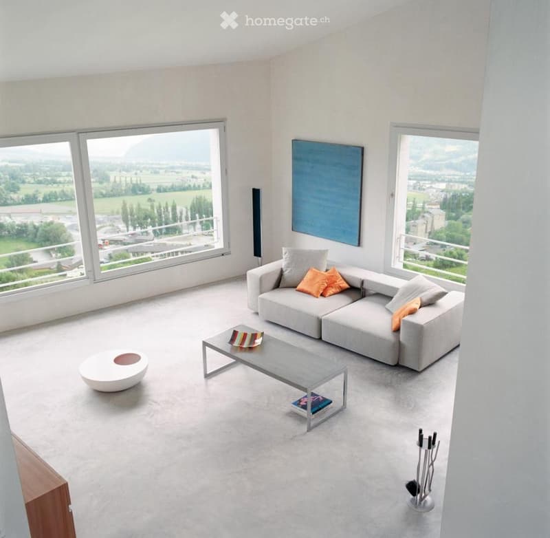 Modern living room with large windows offering a scenic view, featuring a grey sectional sofa with orange cushions, a round white ottoman, a grey coffee table, and a floor lamp.