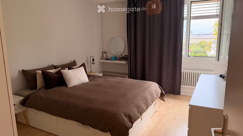 Cozy bedroom with a large bed, dark brown bedding, a desk, a mirror, and a shelf with small items.