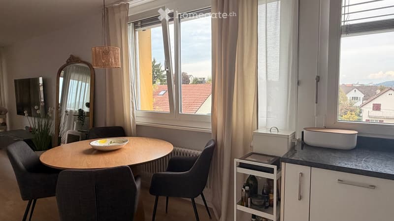 A modern, well-lit room with a round wooden dining table, four chairs, a large mirror, a sink, and a small cabinet. The room features large windows with a view of the city and a hanging lamp.