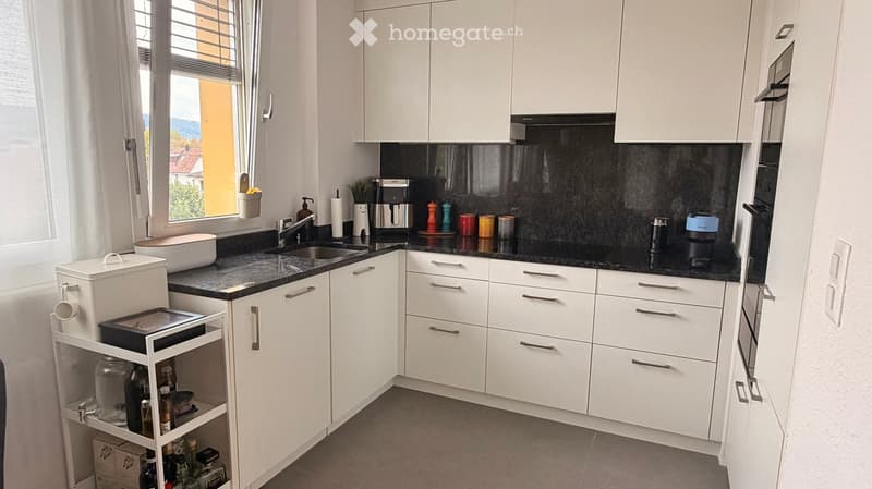 Modern kitchen with white cabinets, black countertops, and various appliances including a coffee maker, sink, and bottle rack. The space features a window with blinds and a small potted plant on the windowsill.