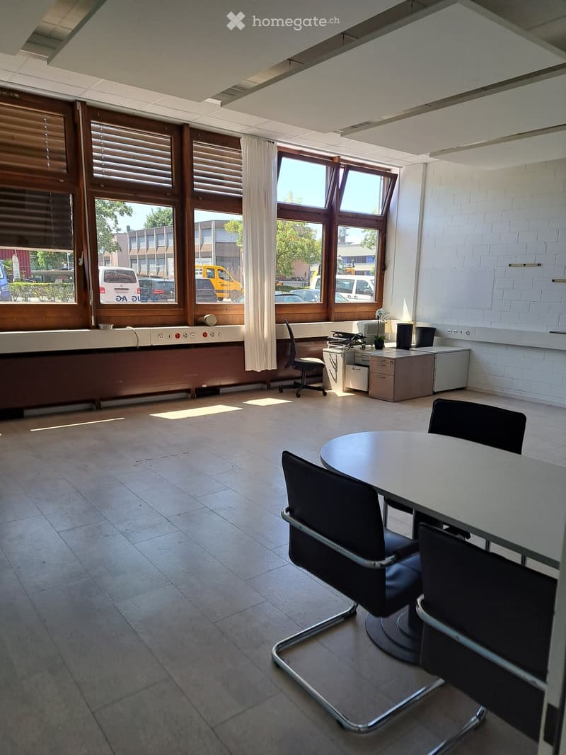 Bright office space with large windows, tiled floor, round table with chairs, office chair, cabinet, printer, and small flower pot.