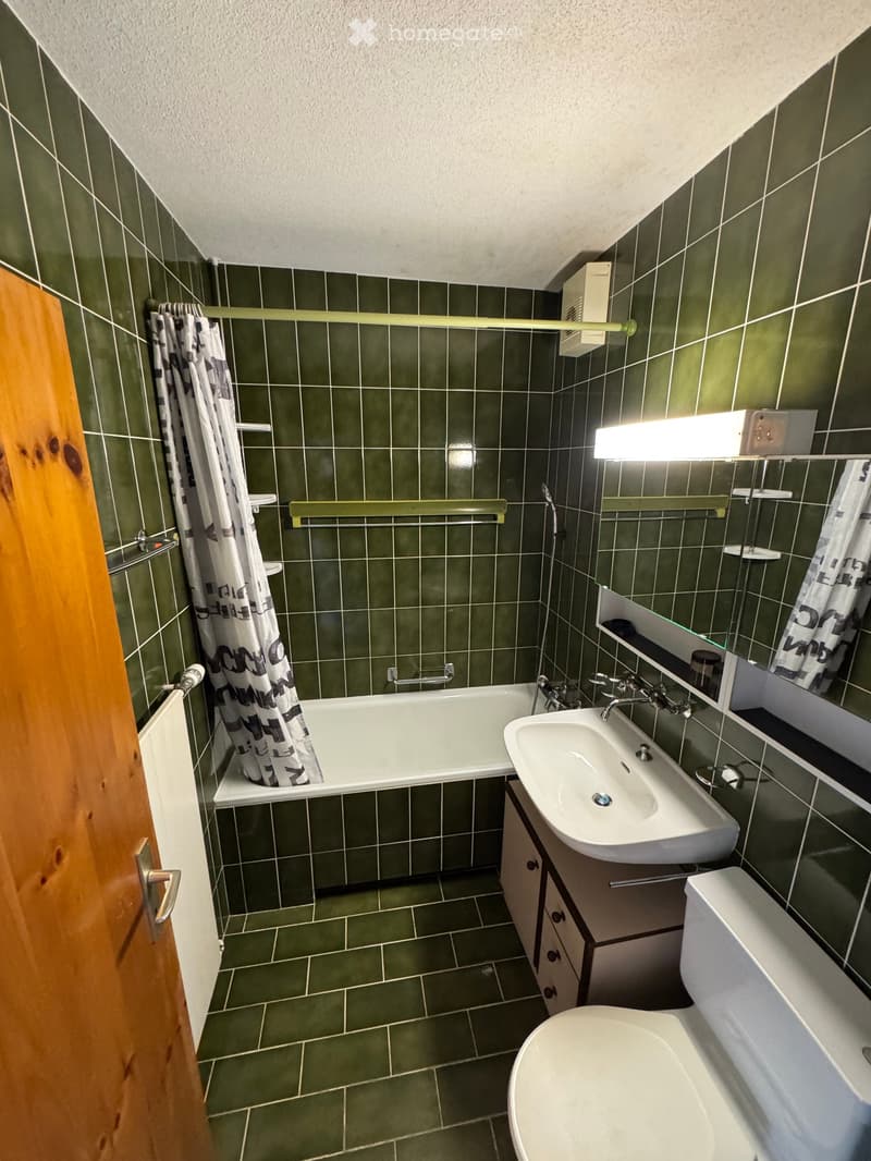 Bathroom with green tiled walls and floor, featuring a bathtub, sink, toilet, mirror, and wooden cabinet.