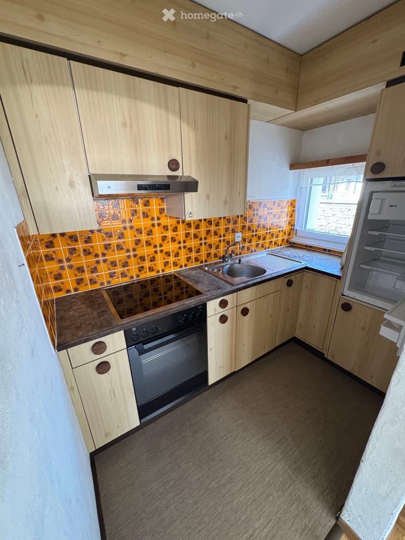 Modern kitchen with light wooden cabinets, stainless steel sink, oven, and refrigerator. Features a tiled backsplash and a window providing natural light.