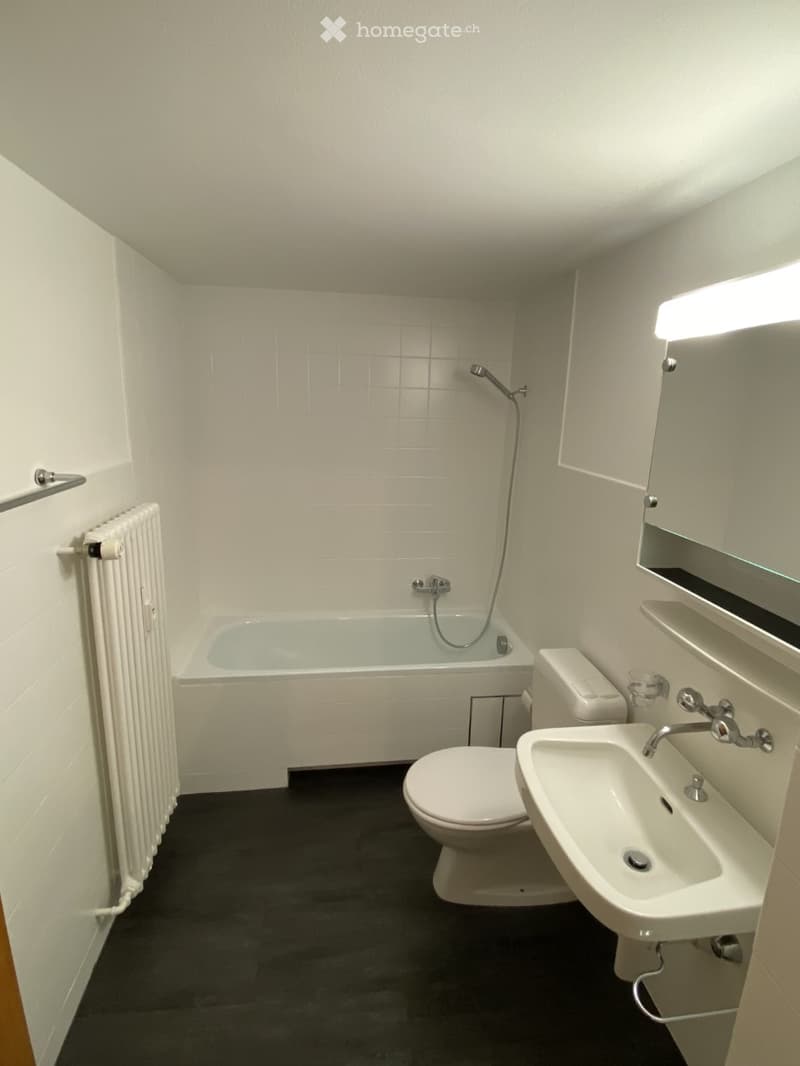 Modern bathroom with white tiles, bathtub, toilet, sink, and a built-in mirror with lighting. Features a dark floor and a radiator on the wall.