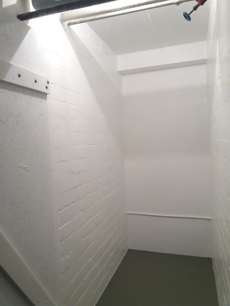 A narrow, white-walled corridor with a slanted ceiling and a small skylight at the top, allowing natural light to enter. The walls are made of white brick, and the floor appears to be concrete.