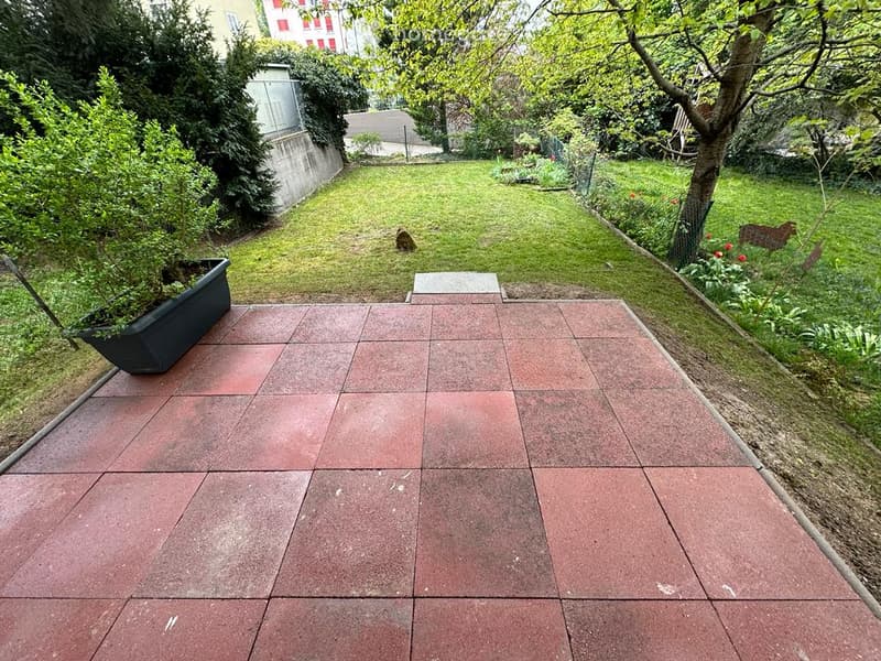 A paved patio area with red tiles leads to a grassy garden, surrounded by trees and shrubs, with a building visible in the background.