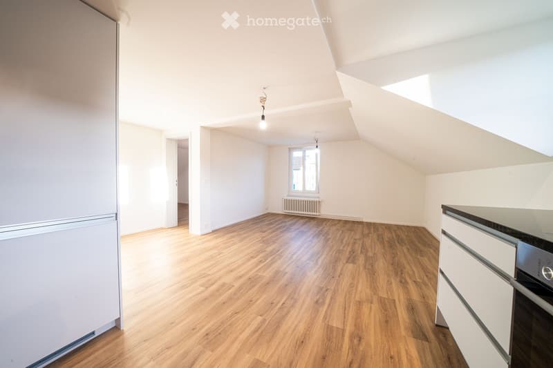 Bright, unfurnished room with wooden flooring, white walls, and modern kitchen cabinets. Features a refrigerator, oven, and a window with natural light.