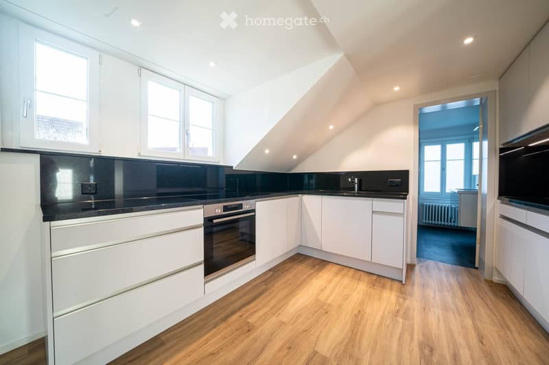 Modern kitchen with white cabinets, black countertops, wooden flooring, and built-in oven. Features large windows and a sleek design.