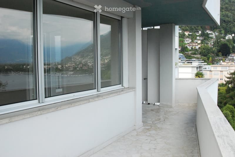 Balcony with large windows offering a scenic view of a town and mountains, with a stone-tiled floor and white walls.