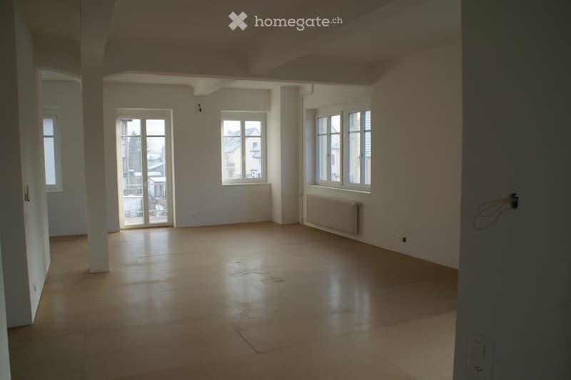 Empty room with tiled floor, white walls, large windows, and a radiator. No furniture or equipment present.