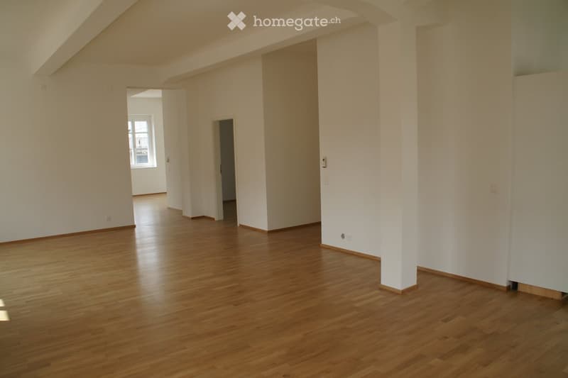 Empty room with wooden flooring and white walls, featuring a doorway leading to another room and a window letting in natural light.