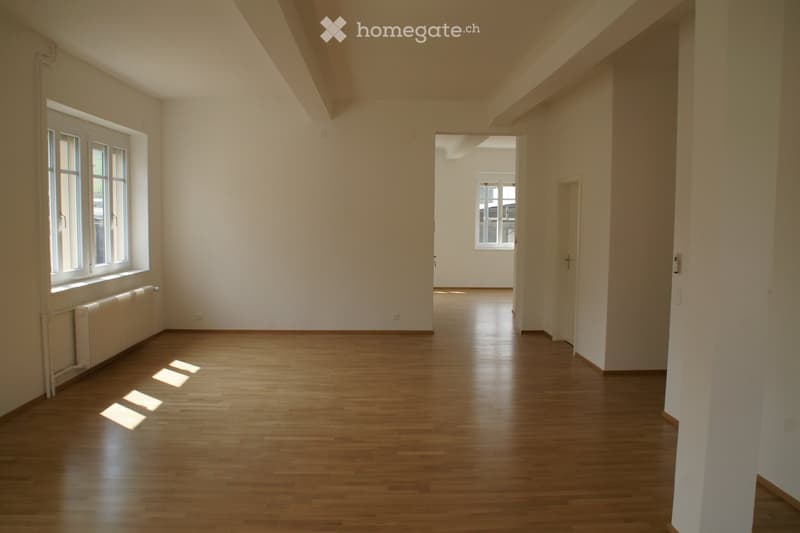 Empty room with wooden flooring, white walls, and large windows allowing natural light. No furniture or personal items are present.
