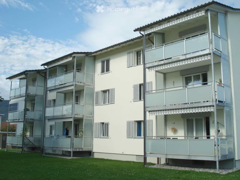 Modern multi-story residential building with balconies and awnings, surrounded by a grassy area.