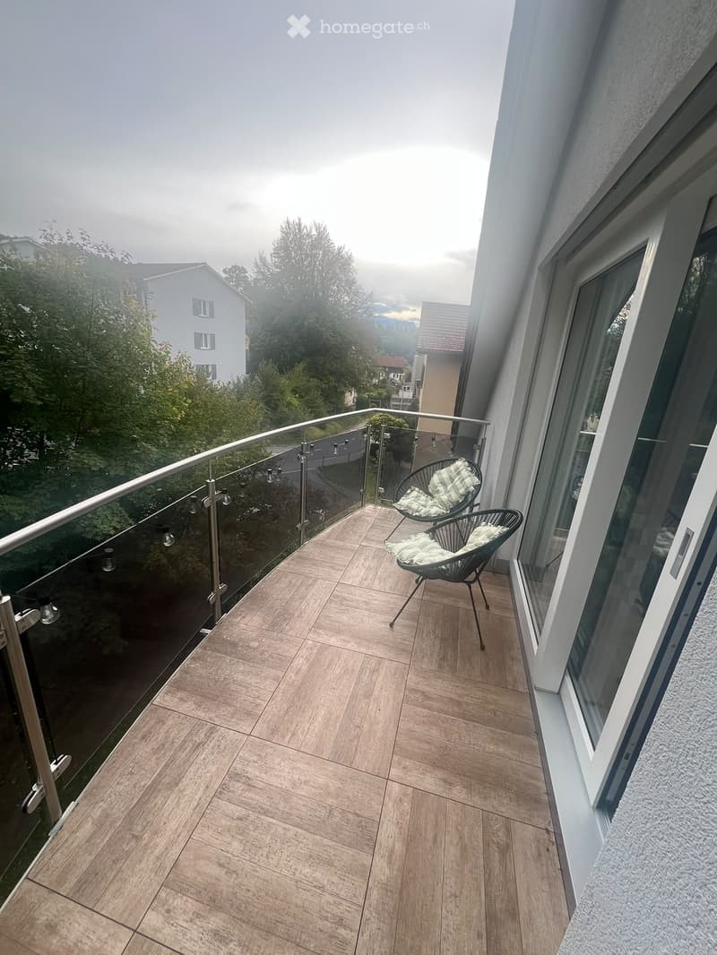 Balcony with wooden floor, glass railing, and two chairs with green cushions.