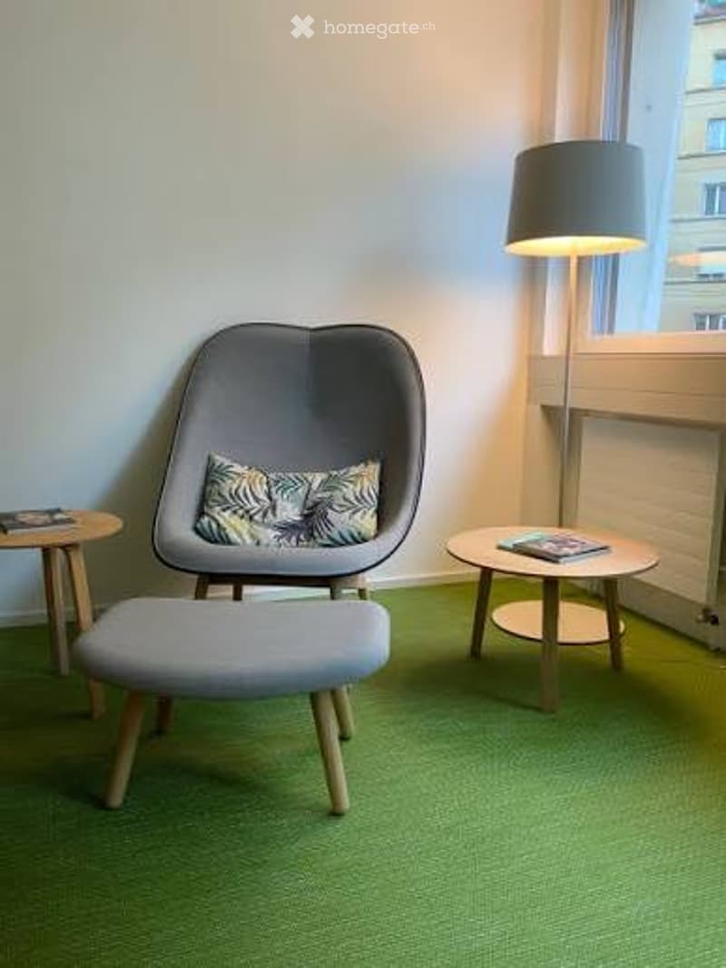 A modern, minimalist room with a grey egg-shaped chair, a matching stool, two small round tables, a floor lamp, and a decorative cushion on the chair.