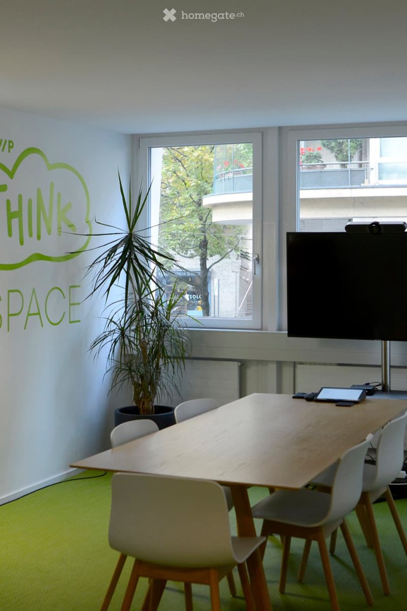 Bright meeting room with a long wooden table surrounded by white chairs, a potted plant, and a large screen on a stand.