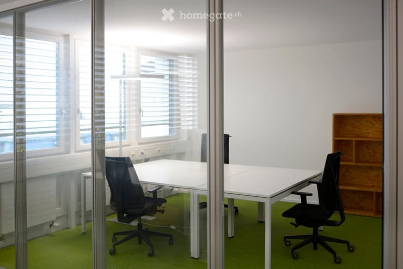 Modern office space with white tables, black office chairs, large windows with blinds, and a wooden shelf unit.