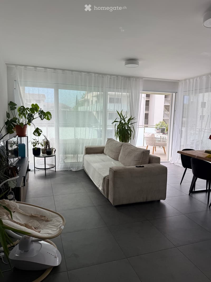 Modern living room with a grey sofa, a small round table, a dining table with chairs, a baby chair, and several potted plants. Large windows with sheer curtains lead to a balcony area with outdoor furniture.