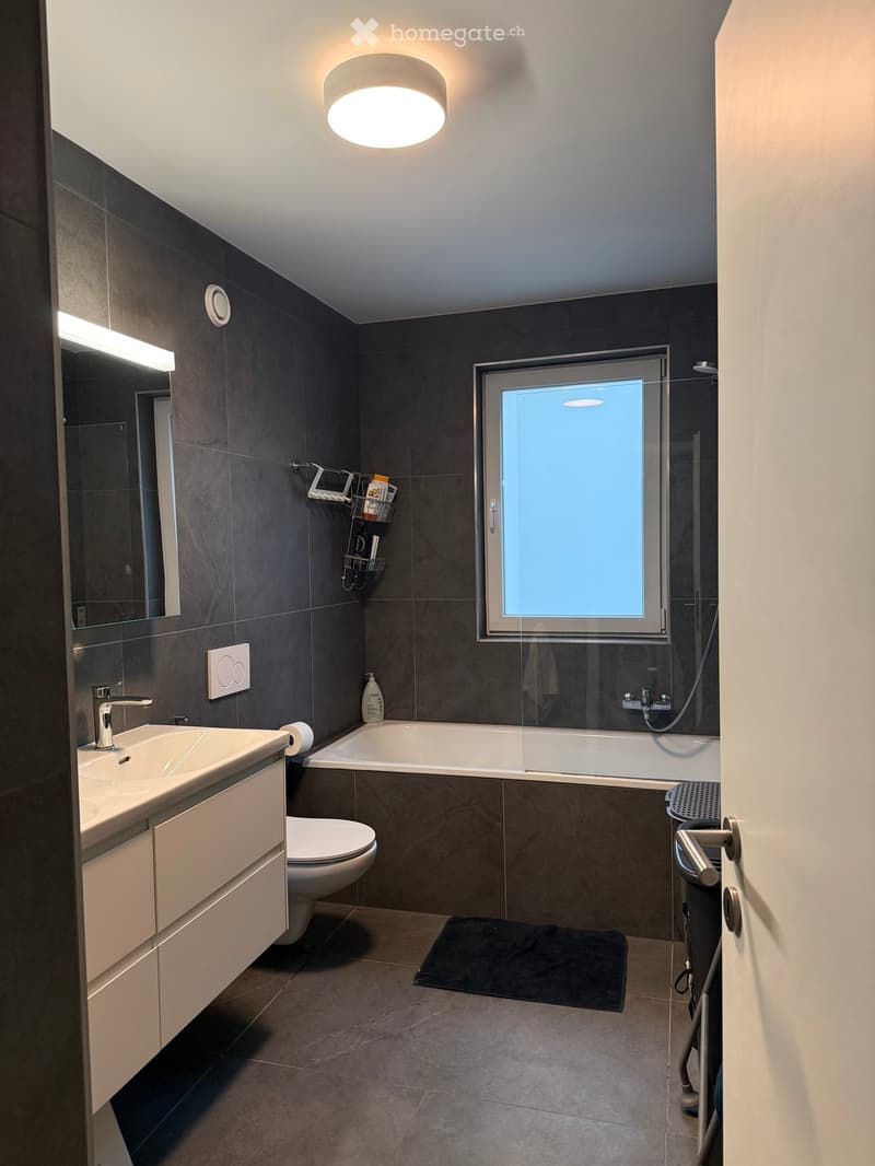 Modern bathroom with dark grey tiles, a white bathtub, a sink with a chrome faucet, a toilet, a mirror, a towel on the floor, and a window with frosted glass.