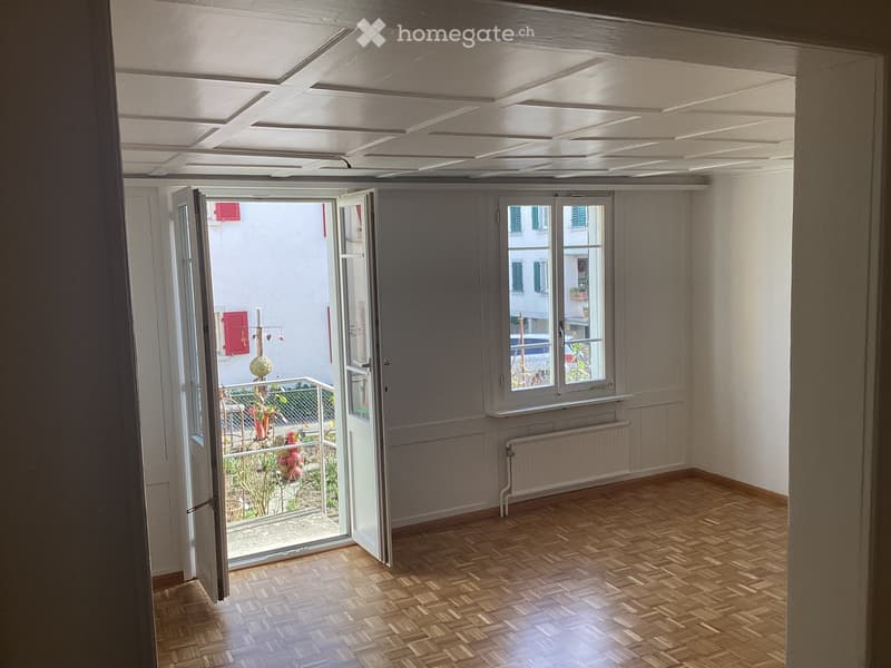 Empty room with wooden parquet flooring, white walls, large window, and open sliding doors leading to a balcony area with plants.