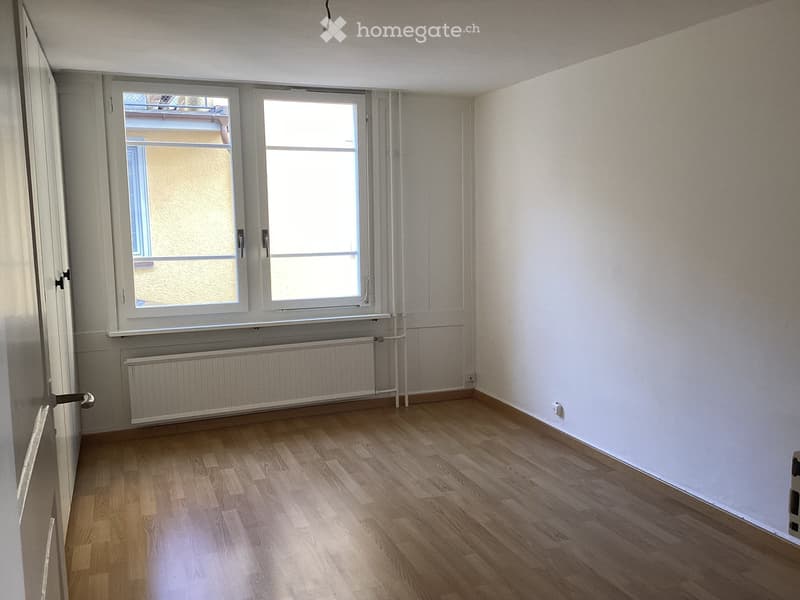 Empty room with wooden flooring, white walls, and large windows with a radiator below them.