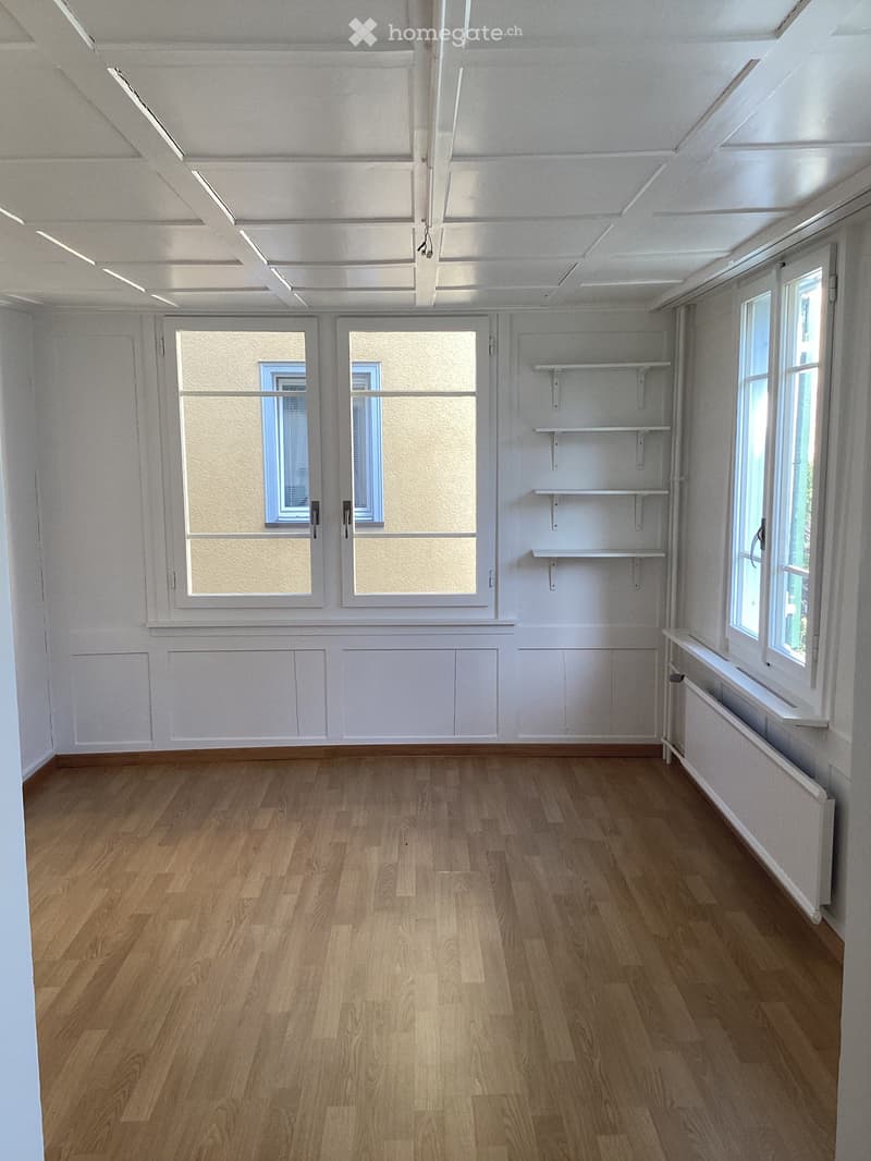 Empty room with wooden flooring, white walls, and large windows with white frames. Shelves are mounted on one wall.