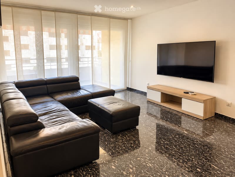 Modern living room with a black leather sectional sofa, ottoman, TV stand, and flat-screen television mounted on the wall. Large windows with sliding glass doors lead to a balcony.