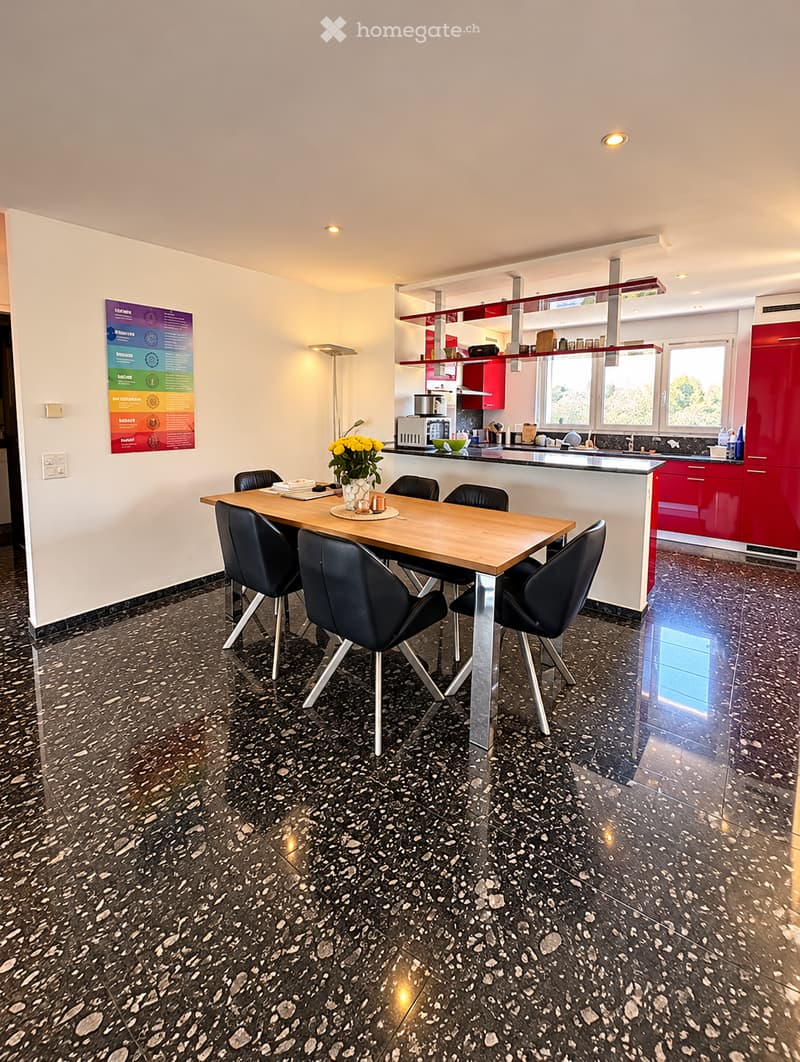 Dining area with a wooden dining table and black chairs, a vase with yellow flowers, and a modern kitchen area with red cabinets, a microwave, and a sink.