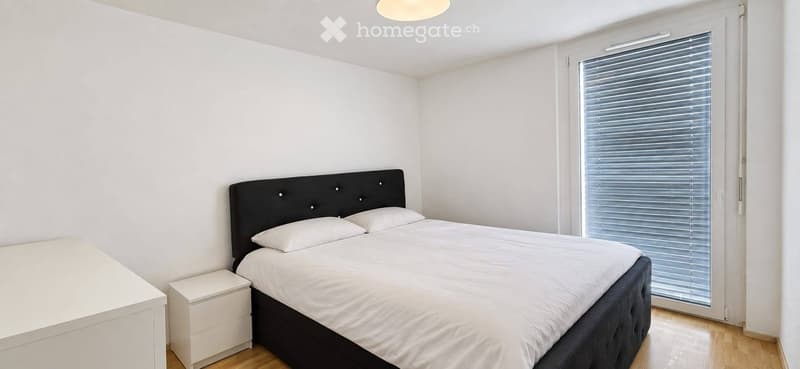 Modern bedroom with a double bed, white bedding, a black headboard, a nightstand, and a large window with blinds.