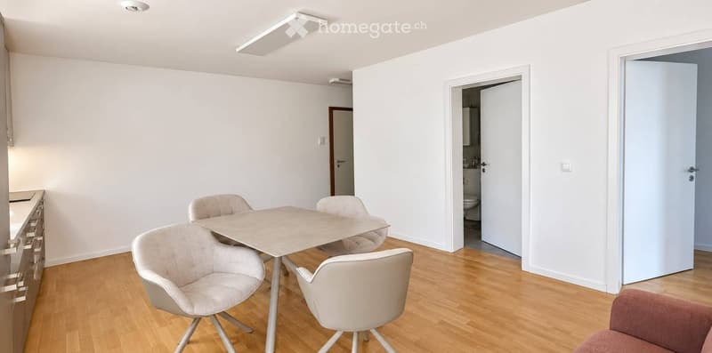 Dining area with a table and four chairs, light-colored walls, wooden flooring, and a cabinet on the left side.