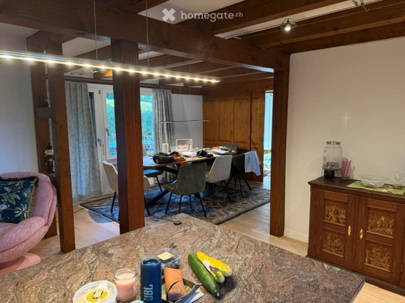 A cozy living and dining area with a pink sofa, a dining table with chairs, a wooden cabinet, and various items on the counter and table.