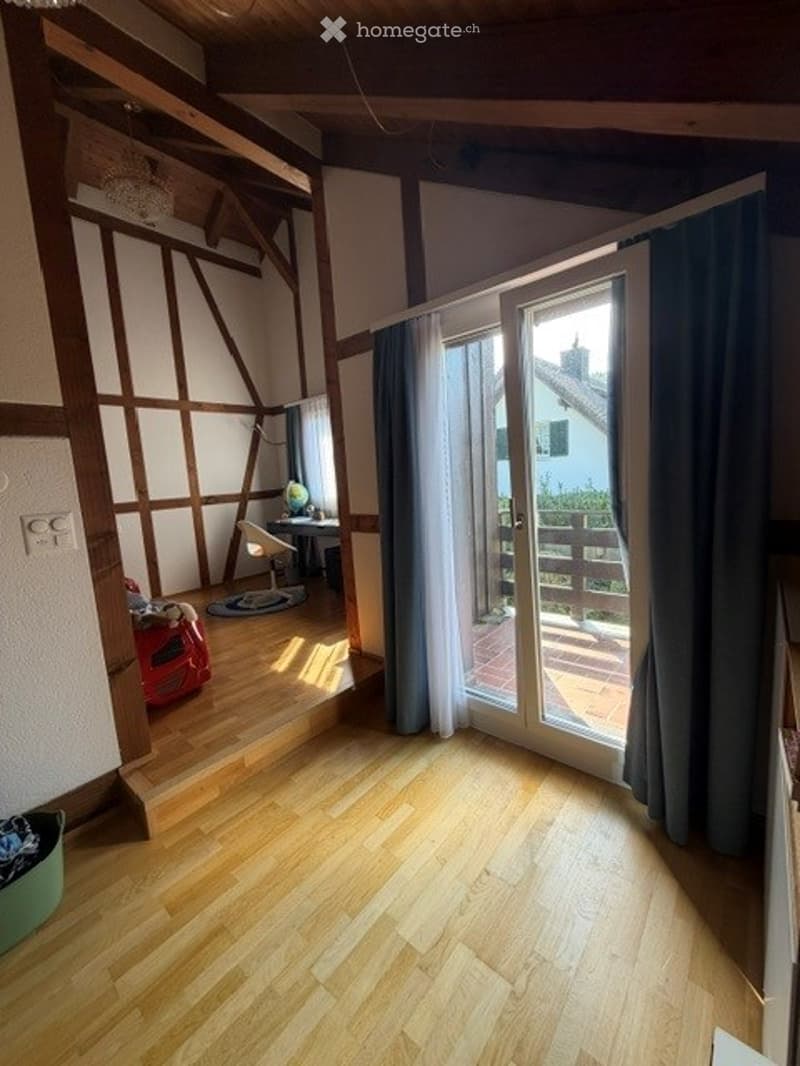 Bright, airy room with wooden flooring, exposed wooden beams, large sliding glass door leading to a balcony, a small seating area with a chair and table, and a globe on a stand.