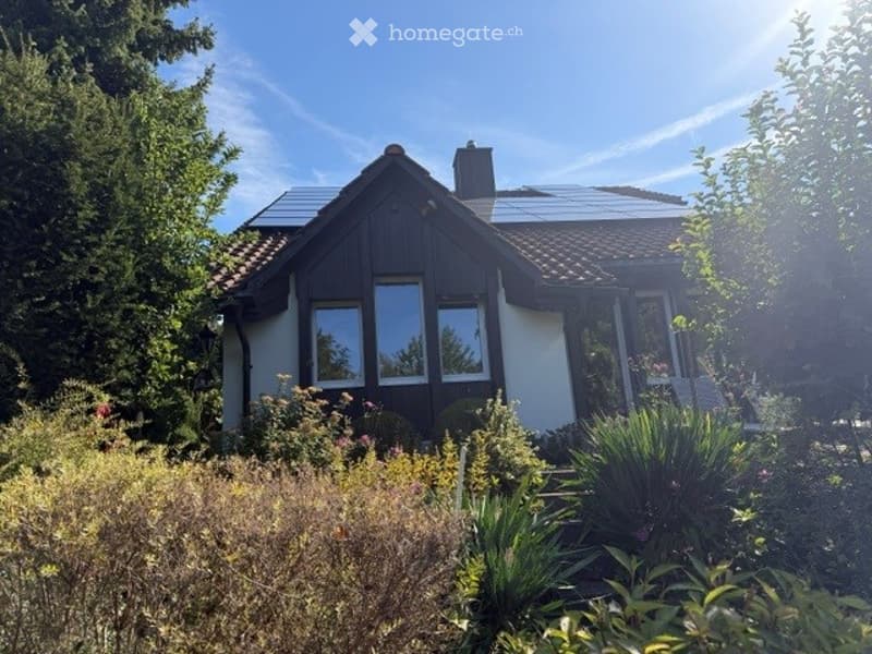 A modern, single-story house with a dark wooden facade and white walls, surrounded by a well-maintained garden with various plants and trees. The roof features solar panels.