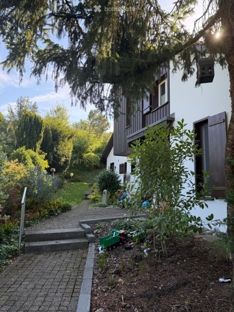 Garden area with a pathway leading to a house, surrounded by greenery and trees. There are small garden decorations and a toy train set visible.